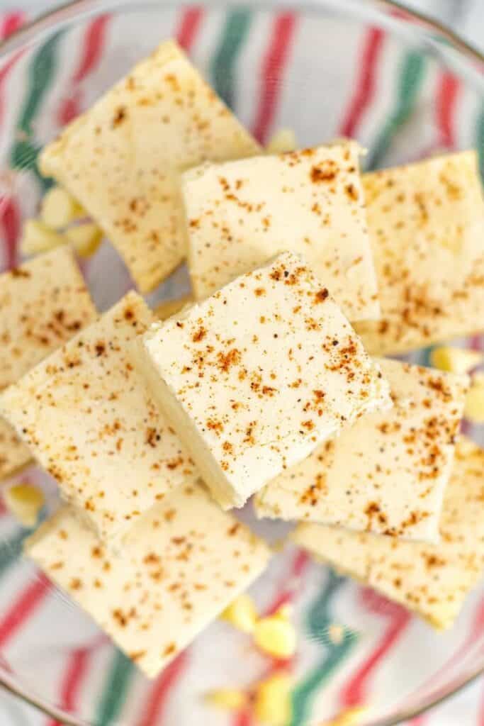 Squares of creamy eggnog fudge sprinkled with nutmeg, stacked on a glass plate. A festive red, green, and white striped cloth peeks through underneath, adding a cheerful holiday backdrop.