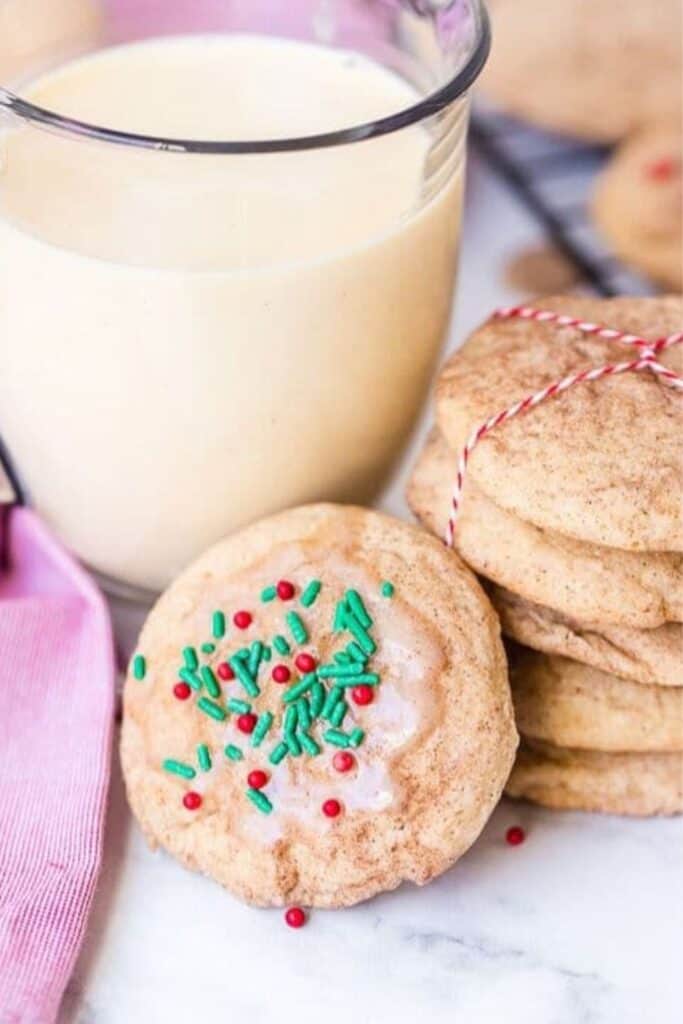 A stack of eggnog snickerdoodle cookies for a cookie exchnage party, some tied with festive string and one decorated with red and green holiday sprinkles, sits beside a glass of creamy eggnog. 