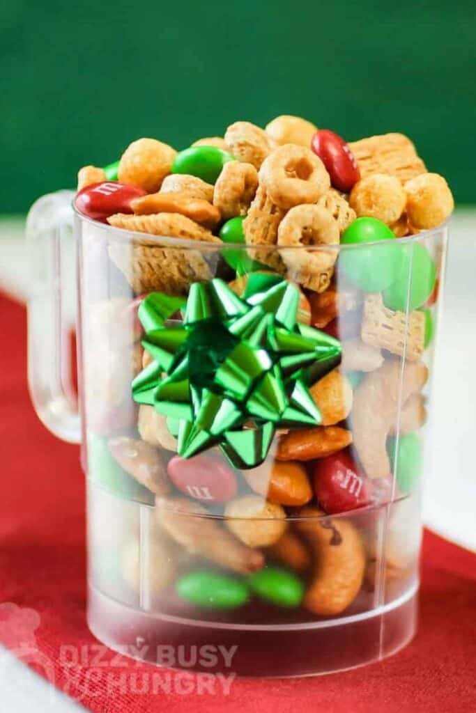 Clear mug of Elf Nibbles snack mix with cherrios, chex cereal, pretzels , M&M's and cashews. 