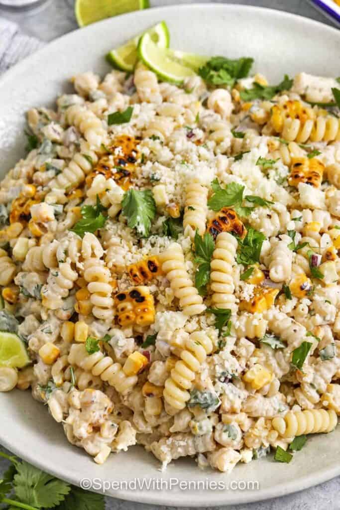 Bowl of elote pasta salad with rotini pasta grilled corn cilantro crumbled cheese and creamy dressing garnished with lime wedges. The Mexican street corn inspired pasta salad is topped with charred corn kernels and fresh herbs.