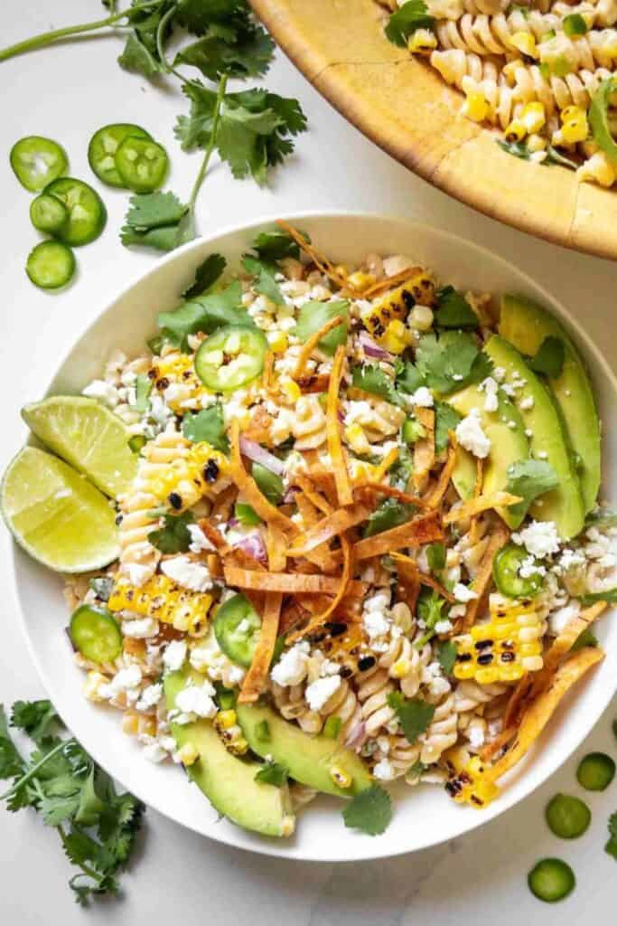 Plate of elote street corn pasta salad with rotini pasta, avocado, feta cheese, jalapeno slices and tortilla strips on top. 