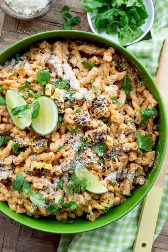 A vibrant green bowl filled with creamy elote pasta salad, featuring grilled corn kernels, corkscrew pasta, chopped cilantro, and a dusting of cotija cheese. Lime wedges and fresh herbs garnish the top, adding a fresh, zesty look.