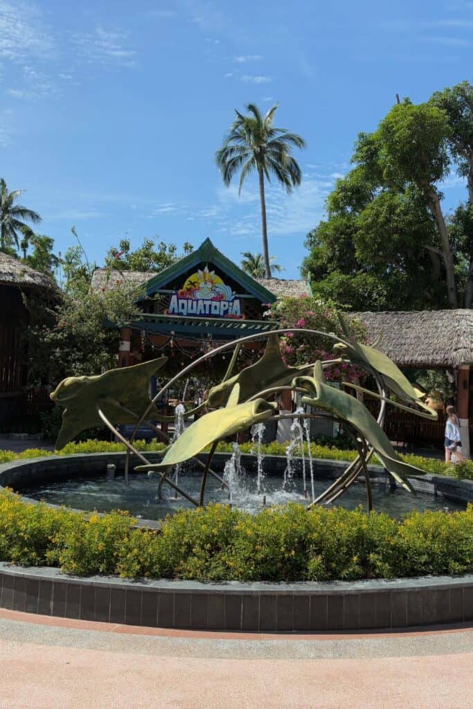A vibrant entrance to Aquatopia Water Park in Phu Quoc, Vietnam, featuring a dynamic fountain with bronze-colored stingray sculptures leaping over spraying jets of water. Palm trees and lush greenery frame the scene under a clear blue sky, with the colorful Aquatopia sign visible above a tropical-style building.