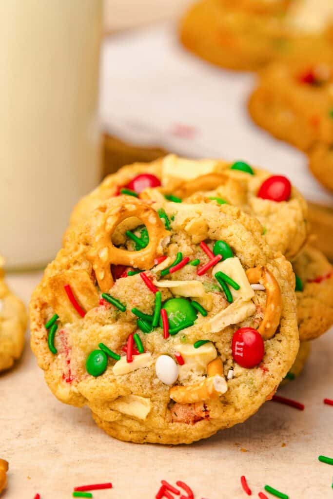 Close-up of a festive Kitchen Sink Christmas cookie packed with red and green M&Ms, pretzel pieces, white chocolate chunks, and red and green sprinkles, with a blurred background of more cookies and a glass of milk.