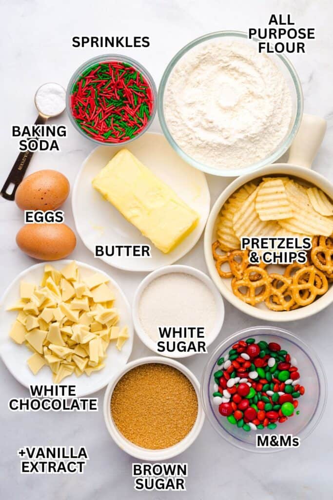 Flat lay of labeled ingredients for Everything Christmas Cookies, including red and green sprinkles, all-purpose flour, baking soda, eggs, butter, pretzels and potato chips, white chocolate chunks, white sugar, brown sugar, M&Ms, and vanilla extract.