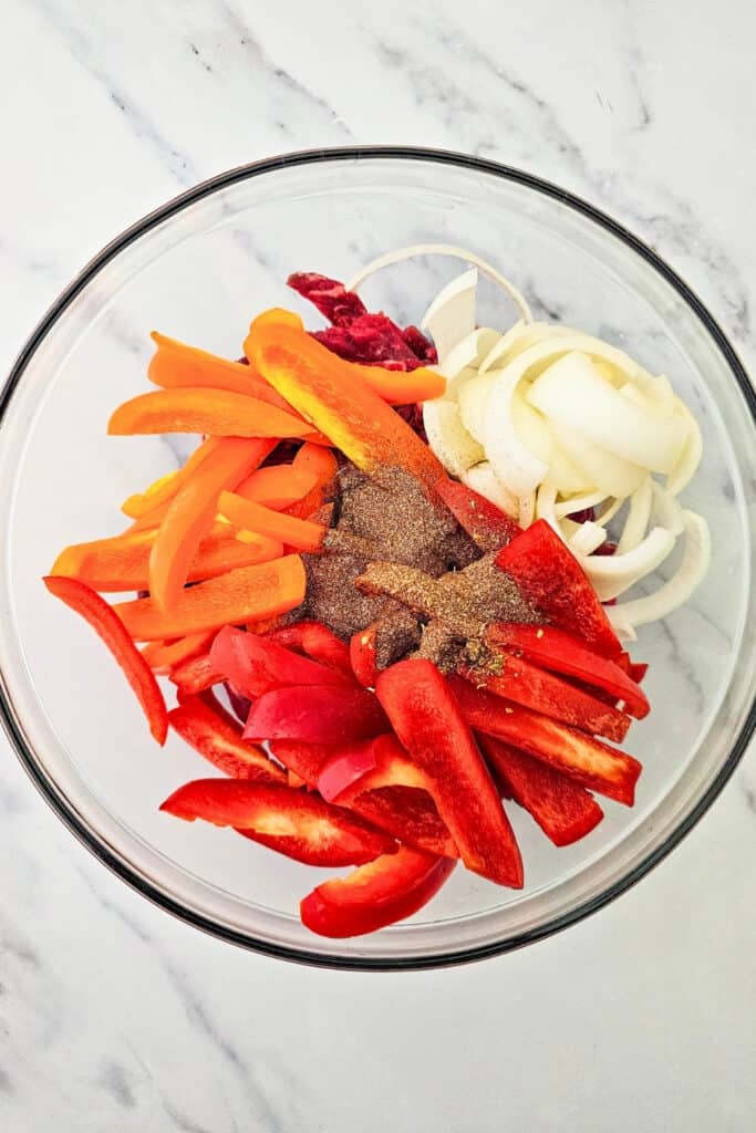 Fajita seasoning is sprinkled over raw steak strips. Sliced red and orange bell peppers and white onions surround the seasoning in a large glass mixing bowl.