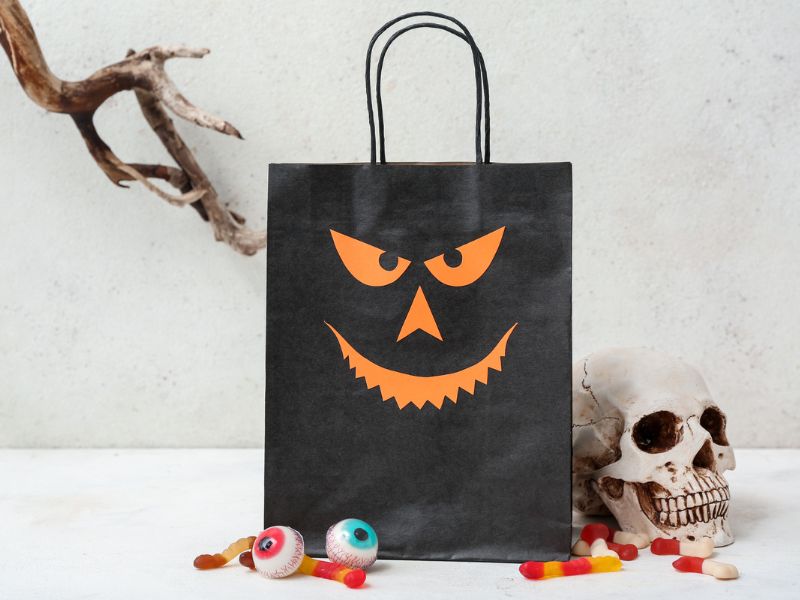 A paper bag with handles decorated with a smiling Jack O' Lantern face. 