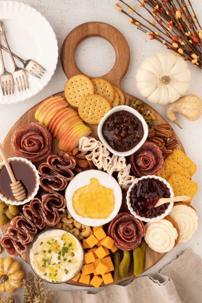 Fall charcuterie board arranged on a wooden pumpkin-shaped tray, featuring cured meats, sliced apples, assorted cheeses, crackers, nuts, fig and berry jams, and mini white pumpkins for decoration. A round brie in the center is topped with pumpkin-shaped jelly.