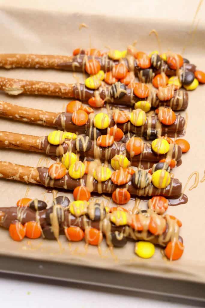 Reese's fall dipped pretzels laid out on parchment paper, coated in melted chocolate and topped with orange, yellow, and brown Reese’s Pieces, then drizzled with creamy peanut butter.