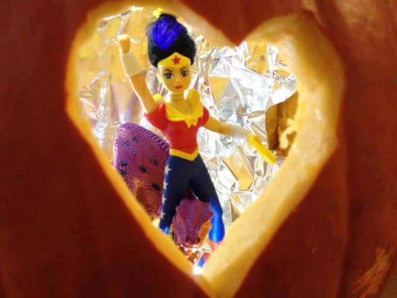 Wonderwoman figurine, riding a toy dinosaur inside a cut out heart shape in a pumpkin.