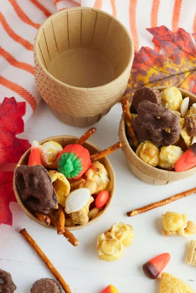 Small brown paper cups filled with a festive fall snack mix, including caramel popcorn, candy corn, chocolate covered pretzels, pretzel sticks, and pumpkin-shaped gummies. Scattered snacks and artificial autumn leaves add to the seasonal, harvest themed presentation.
