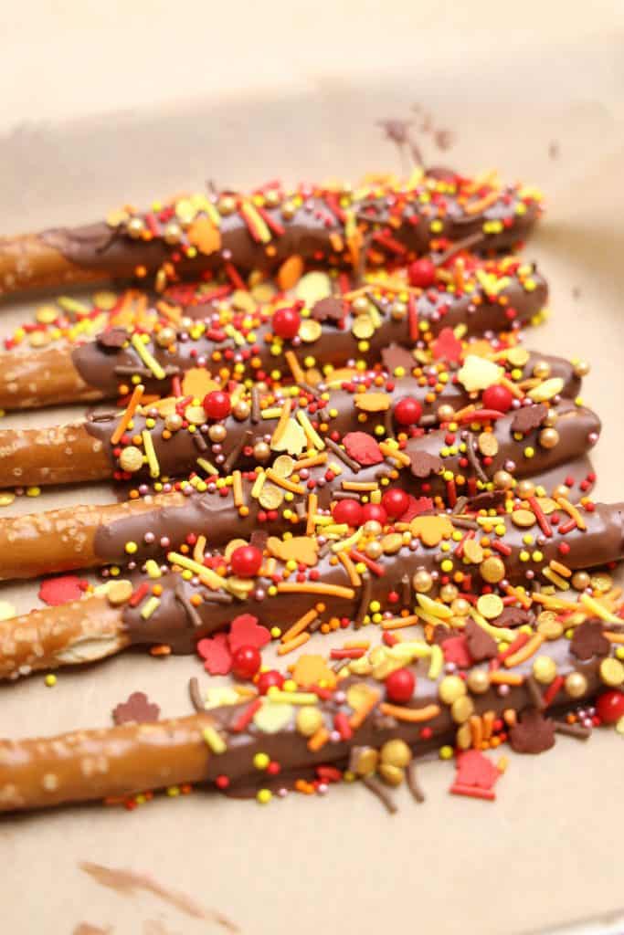 Fall Themed Chocolate Dipped Pretzel Rods arranged on parchment paper, coated in milk chocolate and topped with colorful autumn sprinkles including leaves, red and gold balls, and orange and yellow jimmies.