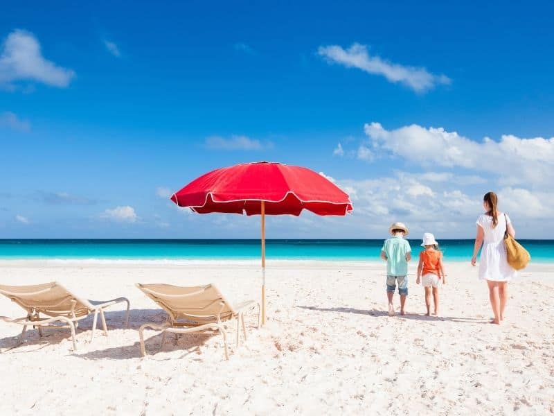 Mother, young boy and girl on beach beside two chairs and umbrella. Ocean in background. 