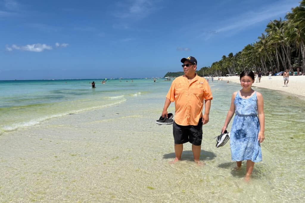 A dad and tween daughter walking in ankle deep turquoise water on a sandy tropical beach with Crocs and runnung shoes in hand. 