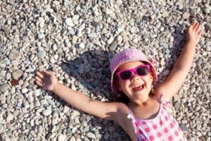 young girl lying on beach pebbles wearing bathing suit, sun hat and sunglasses smiling. USA spring break destinations