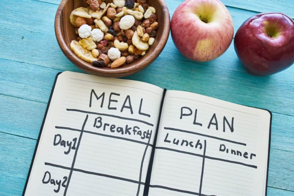 Notebook open to meal planning, with two red apples and and a wooden bowl of trail mix beside it.