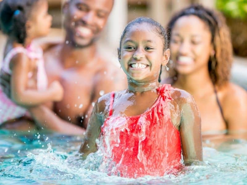 Family Vacation. Young girl with big smile jumping out of water in pool. Dad and mom behind.