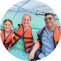 A smiling family of three wearing orange life jackets sits on a boat with turquoise water and distant boats in the background. The cloudy sky suggests overcast weather during a scenic outing.