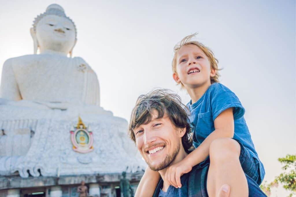 Young boy on top of dad's shoulders smiling in front of a big Buddha statue in Phuket, Thailand