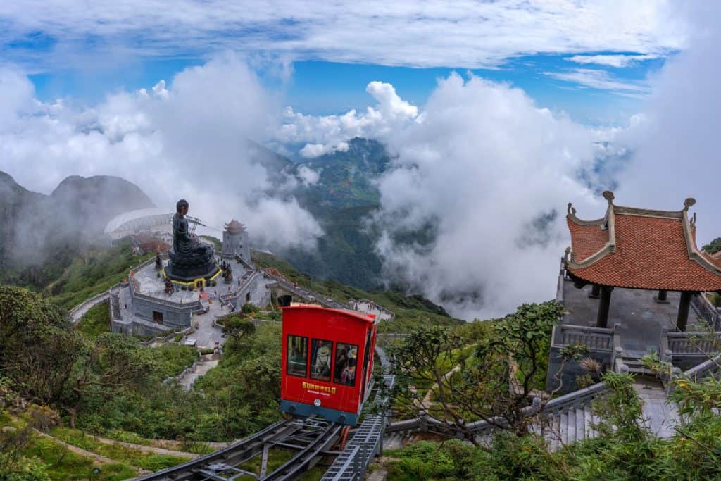 A red funicular railway car ascends through the clouds toward the summit of Fansipan Mountain in Vietnam, passing by a massive seated Buddha statue surrounded by a stone courtyard and temple structures. The scene is set high above the clouds, with dramatic views of forested valleys, mountain peaks, and traditional Vietnamese architecture.