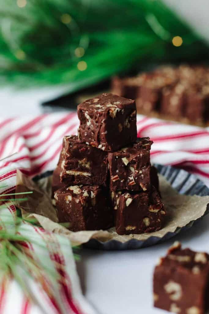 Stacked cubes of classic fantasy fudge filled with chopped pecans, displayed on a fluted dish lined with parchment paper. A festive red and white striped cloth and blurred green garland in the background add cozy holiday charm.