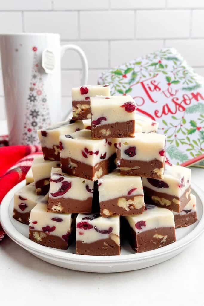 A plate stacked with square pieces of layered Christmas fudge made with white and milk chocolate, studded with dried cranberries and chopped nuts. In the background, a festive holiday mug and a sign reading “’Tis the Season."
