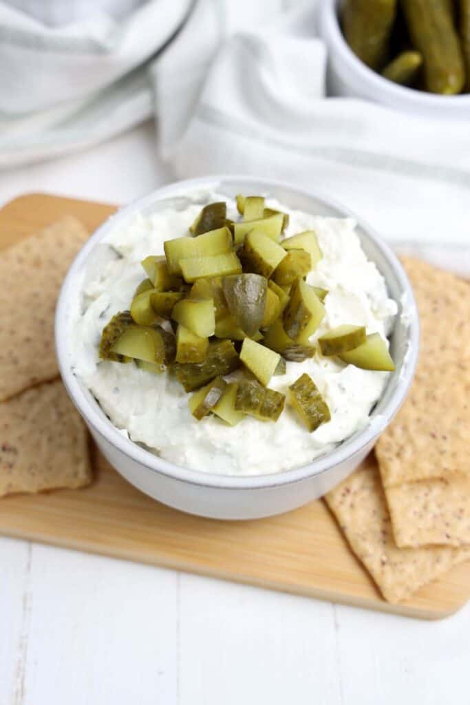 An overhead view of a bowl of festive and creamy dill pickle dip on a wooden board, surrounded by crackers. 