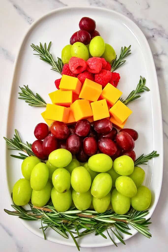 A festive fruit platter arranged in the shape of a Christmas tree with colorful layers of green and red grapes, fresh raspberries, cheddar cheese cubes, and rosemary branches on a white oval serving tray.