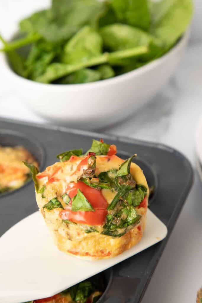 Feta spinach egg muffin topped with wilted spinach and diced red pepper sitting on a small plate with a muffin tin in the background for a savory breakfast. 