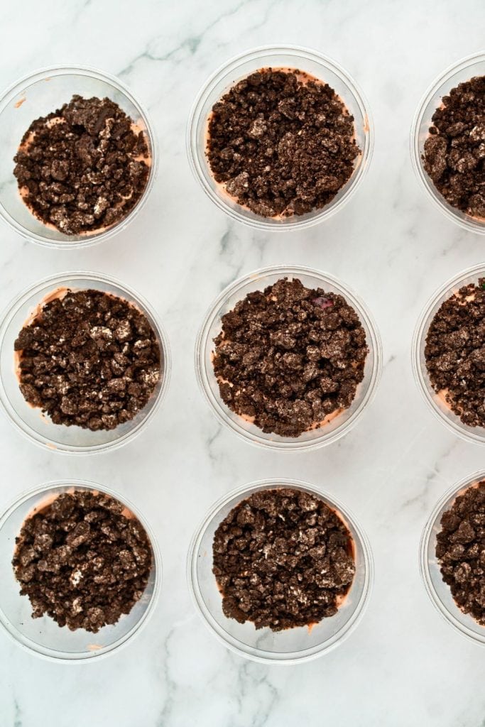 Top-down view of clear plastic cups filled with layers of chocolate pudding mixture and topped with crushed chocolate cookies, arranged neatly on a marble surface.
