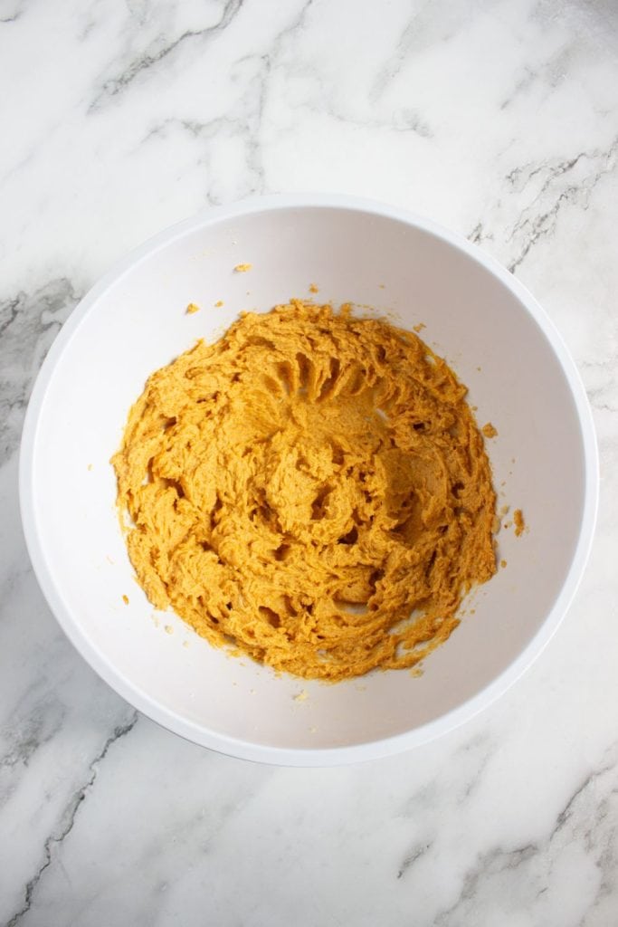 Bowl of smooth, orange pumpkin spice cookie dough fully mixed and ready to chill or shape. The dough sits in a white mixing bowl on a marble countertop, rich with warm fall color.