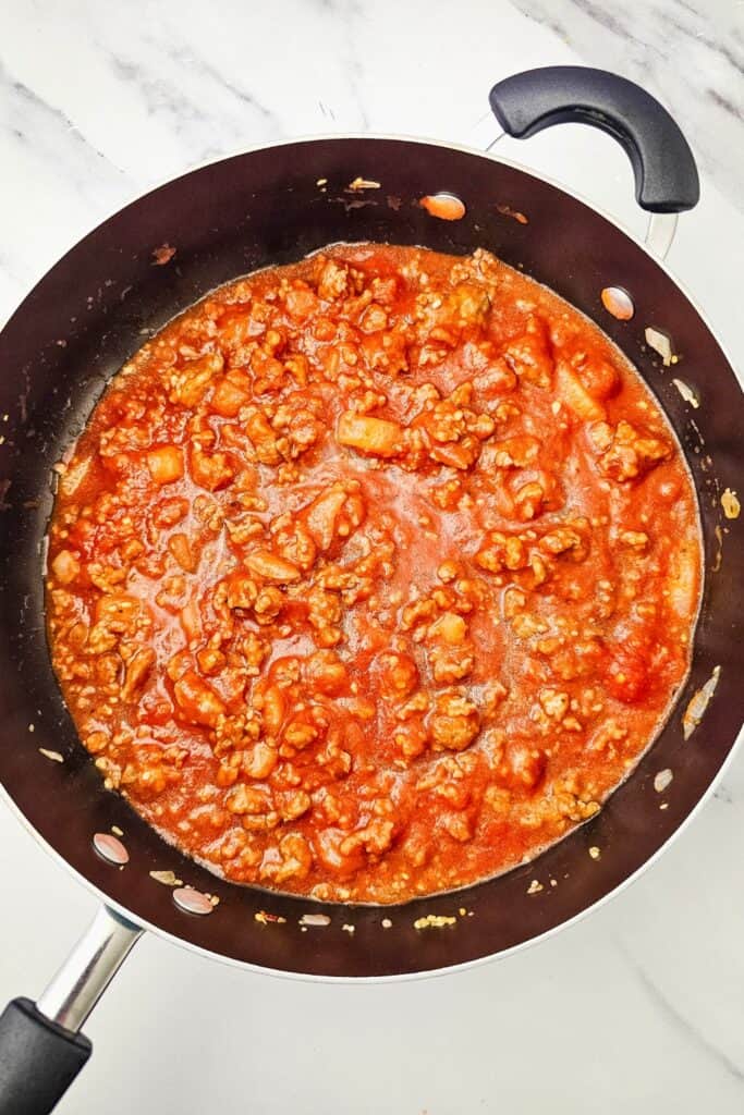 Overhead view of Italian sausage meat sauce simmering in a large skillet with tomato sauce and softened onions. The rich red sauce will be used to assemble the lasagna.