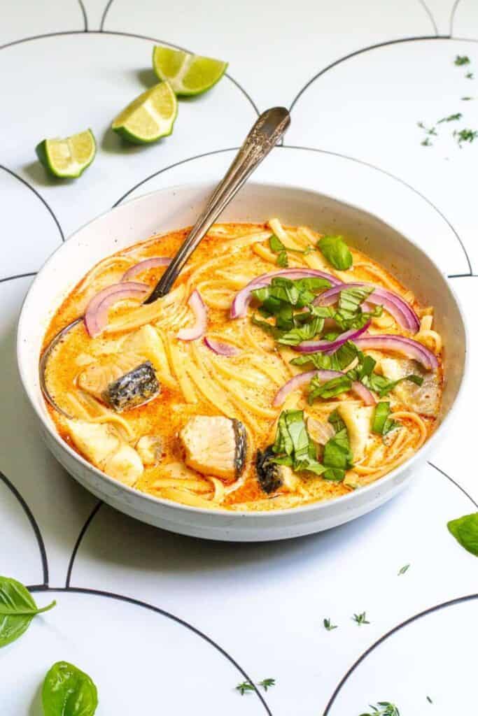 Fish coconut curry soup with chunks of fish and noodles is served in a creamy orange coconut broth. Fresh basil red onion and lime add colorful garnishes to the soup.