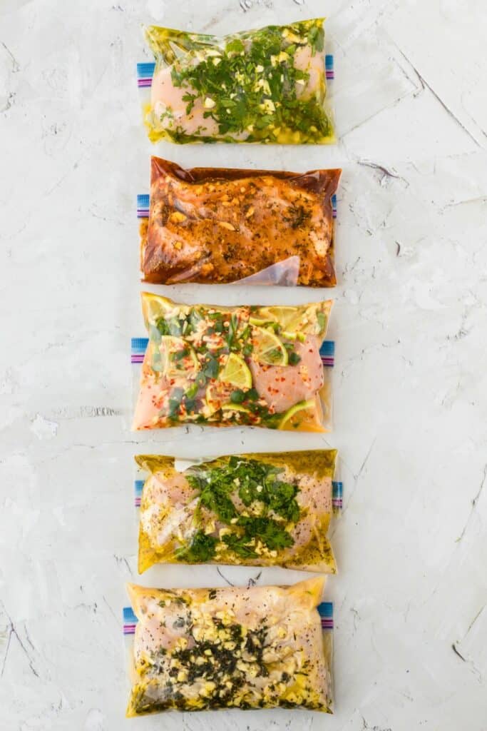 Five raw chicken breasts sealed in zip-top bags, each marinating in different flavorful mixtures with visible herbs, garlic, citrus slices, and spices.
