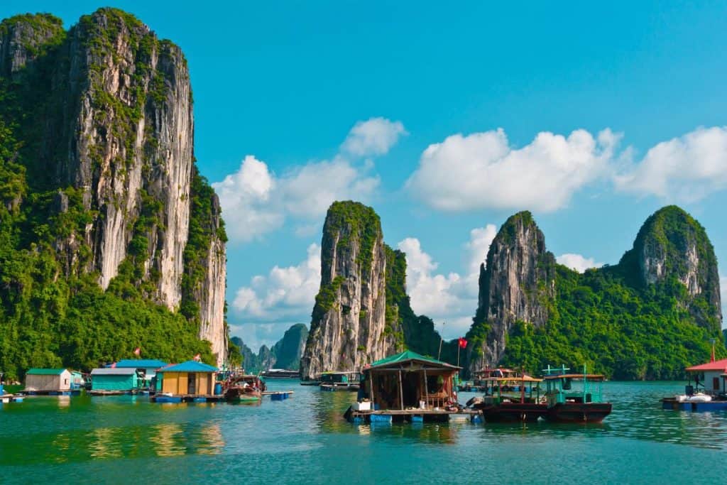 Towering limestone karsts covered in lush greenery rise dramatically from the emerald waters of Ha Long Bay in Vietnam. Colorful floating houses and boats cluster in the foreground, creating a picturesque scene under a bright blue sky with scattered clouds.