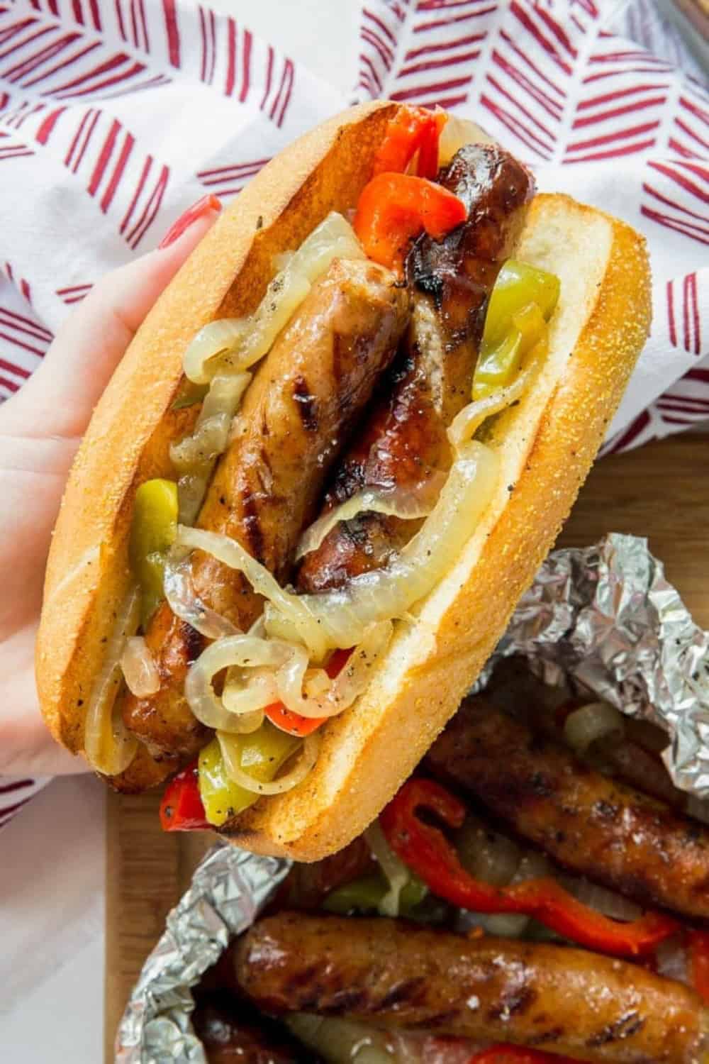 Grilled sausages tucked into a toasted roll and topped with saut&eacute;ed onions and sliced red and green peppers. The sandwich is held above a tray of foil packet sausages and vegetables, showing the charred sausage and tender peppers inside the bun.