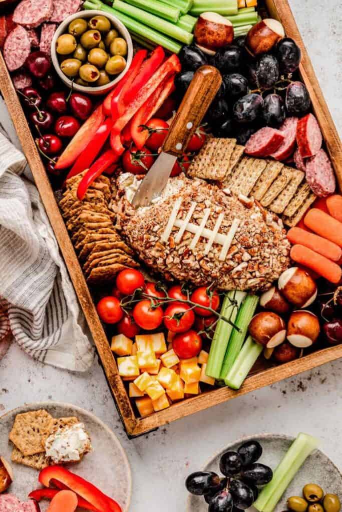 A vibrant wooden charcuterie board styled as an easy football game day appetizer, featuring a football-shaped cheese ball topped with almond coating and white cheese laces, surrounded by crackers, cherry tomatoes, celery, red peppers, salami, olives, baby carrots, grapes, pretzel rolls, and cheese cubes.