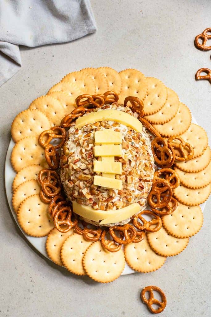 Football-shaped cheese ball coated in chopped pecans and topped with cheddar cheese laces, surrounded by round buttery crackers and mini pretzels. A fun tailgate food arranged on a white plate against a neutral background.