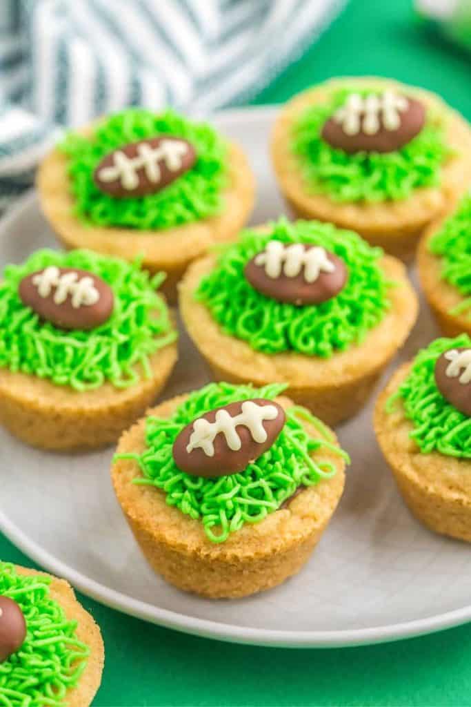 Football cookie cups decorated with green frosting to resemble grass and topped with chocolate footballs piped with white icing laces. The cookie cups are arranged on a white plate over a green background.