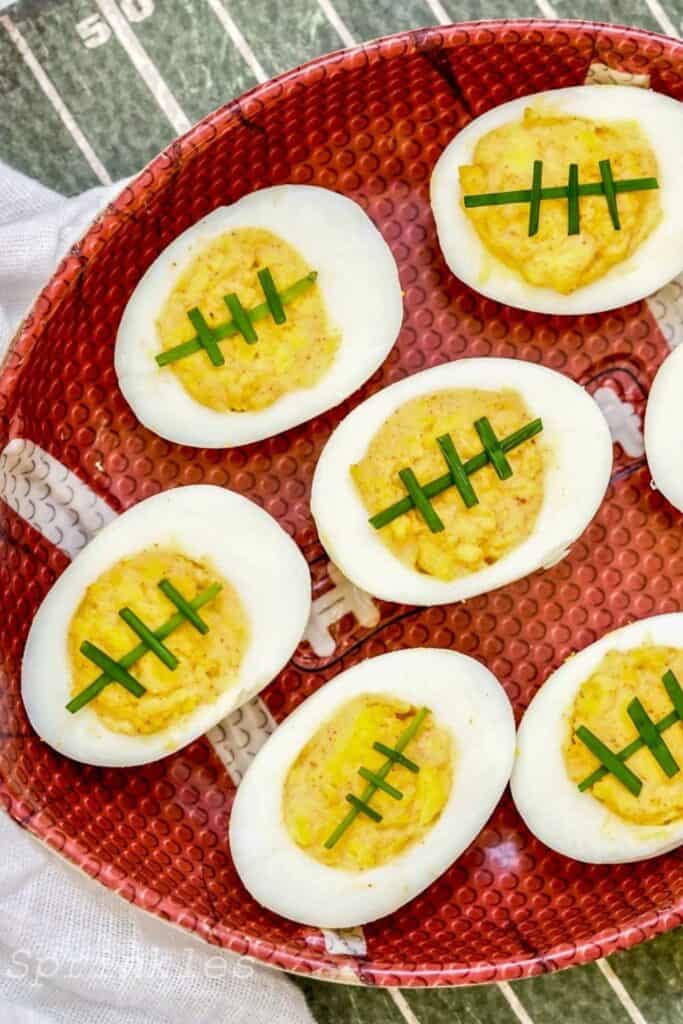 Deviled eggs styled like footballs, with the yolk filling topped with thin slices of chives to mimic football laces, arranged on a red-brown football-shaped platter set over a field-inspired background.