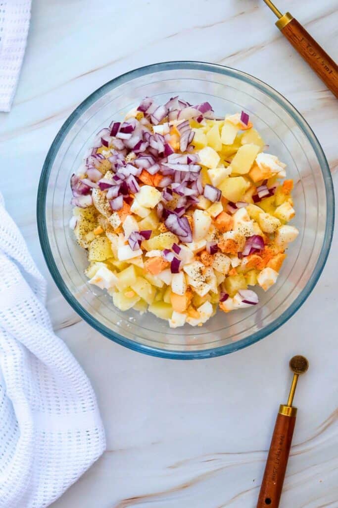 Overhead process shot of a clear bowl of chopped eggs, red onion, and cubed potatoes. 