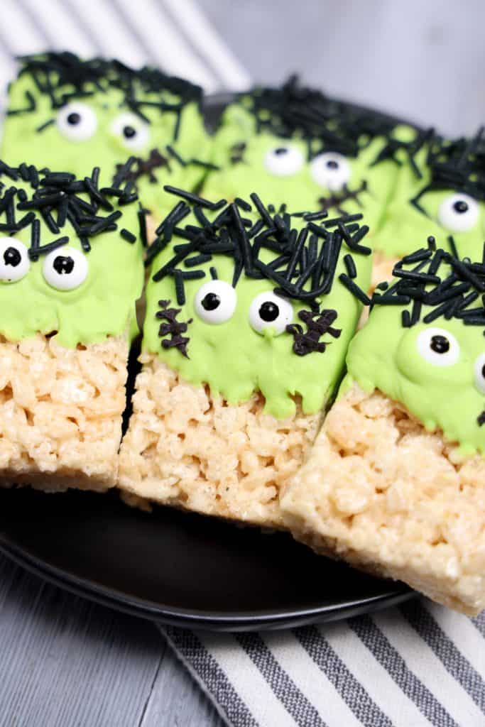 Close-up of Frankenstein-themed Rice Krispie treats decorated with green frosting, candy eyes, and black sprinkles for hair and stitches, arranged on a black plate over a striped cloth.