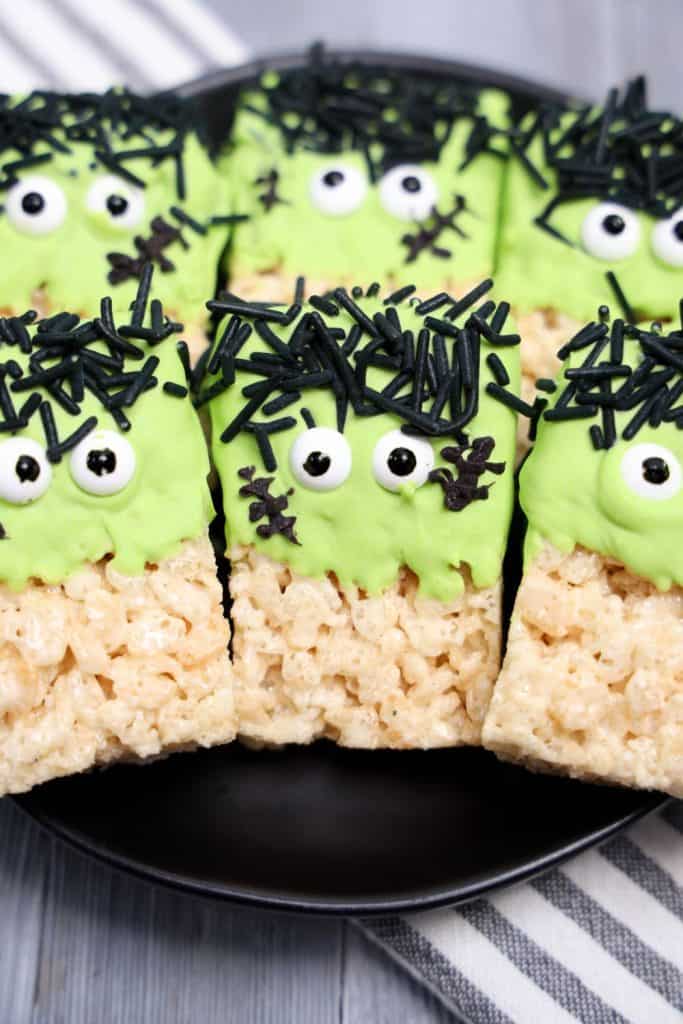 Finished Frankenstein Rice Krispie treats displayed on a black plate, each decorated with green candy coating, candy eyes, black jimmies for hair, and chocolate stitch marks to resemble the classic Halloween monster. The plate rests on a striped cloth over a wooden surface.