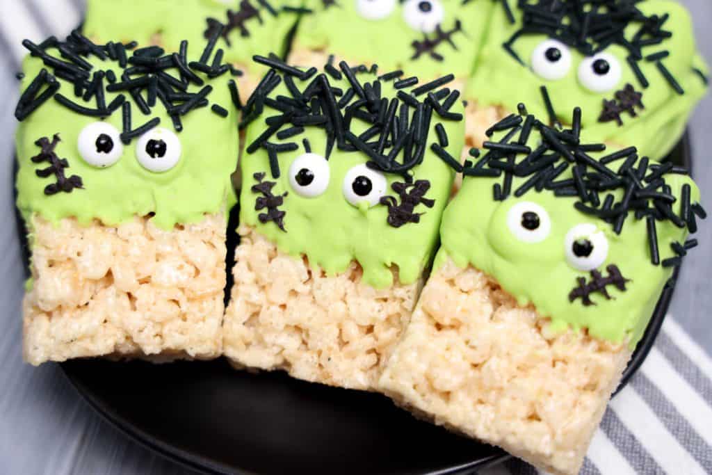 Halloween party snacks featuring Frankenstein Rice Krispie treats decorated with green frosting, candy eyes, and black sprinkles for hair and stitches. The spooky-themed treats are neatly arranged on a black plate over a striped cloth, perfect for a festive gathering.