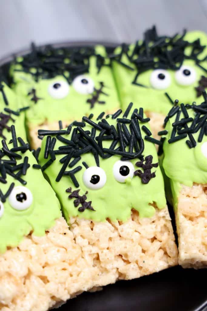 Close-up of Frankenstein Rice Krispie treats decorated with green candy coating, candy eyes, black jimmies for hair, and chocolate stitches. The treats are arranged tightly on a black plate, showing their playful Halloween monster faces.