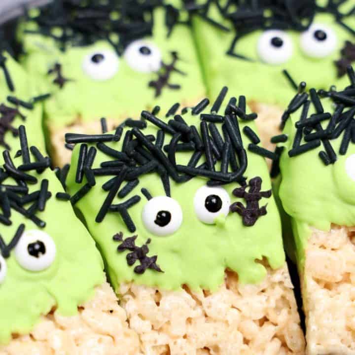 Close-up of Frankenstein Rice Krispie treats decorated with green candy coating, candy eyes, black jimmies for hair, and chocolate stitches. The treats are arranged tightly on a black plate, showing their playful Halloween monster faces.