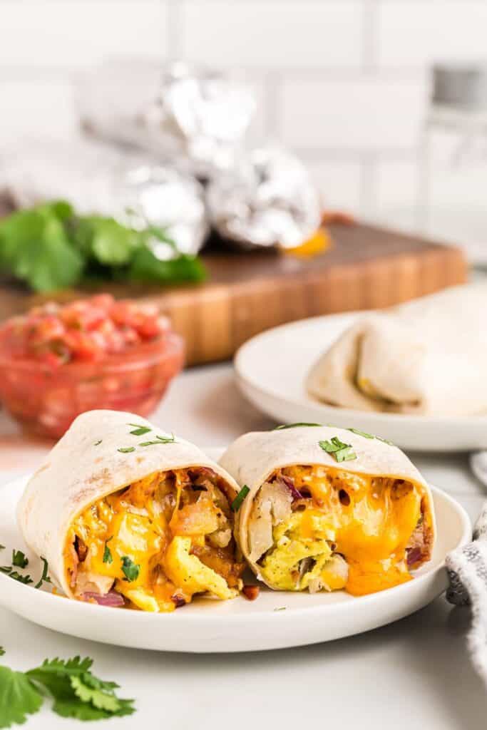 Freezer breakfast burritos sliced in half to reveal fluffy scrambled eggs, crispy potatoes, melted cheddar cheese, and bits of ham wrapped in a soft flour tortilla. The burritos sit on a white plate with fresh cilantro sprinkled on top, and a bowl of salsa in the background for serving.