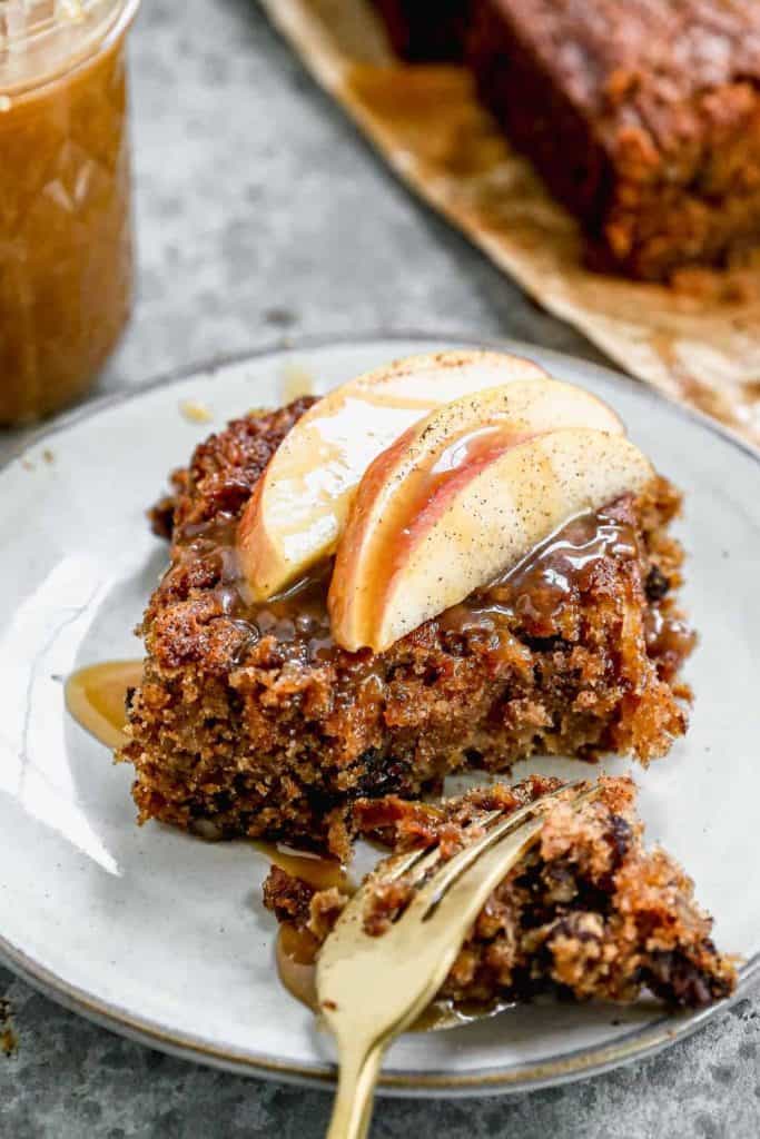 A moist slice of fresh apple cake topped with caramel sauce and thinly sliced apples, served on a white plate with a gold fork. The dense, spiced crumb is studded with fruit and nuts, making it a cozy dessert for fall.