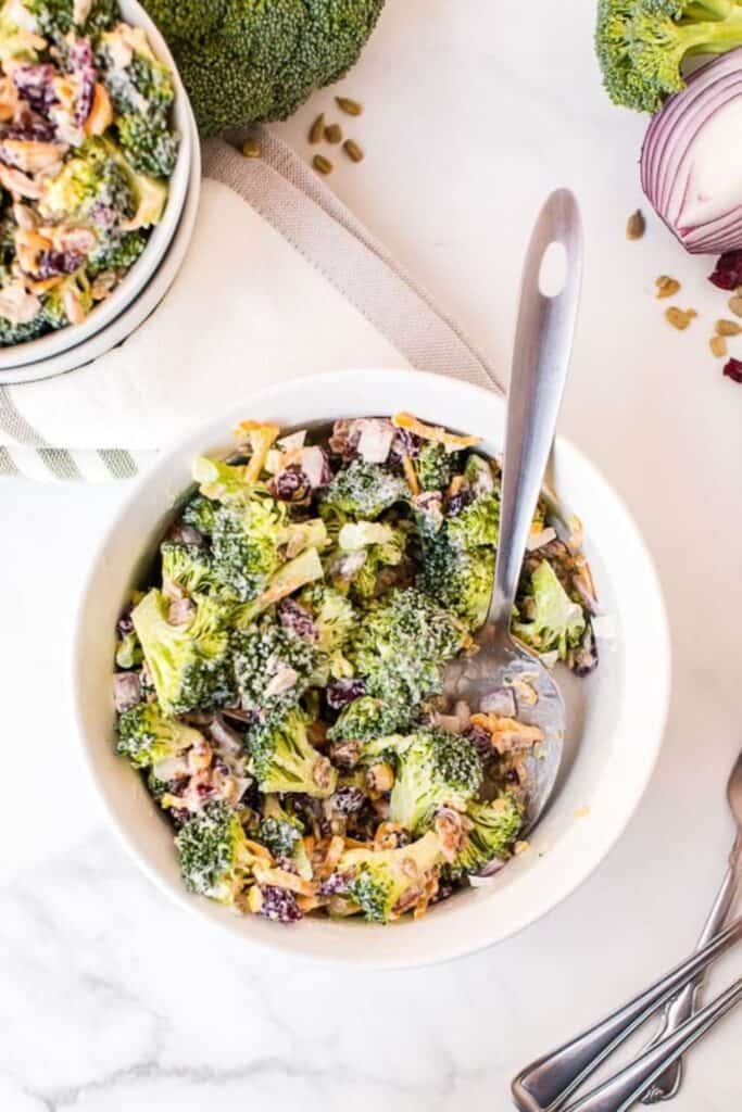 Creamy broccoli salad in a white bowl, made with fresh broccoli florets, shredded carrots, red onion, sunflower seeds, and dried cranberries, all tossed in a creamy dressing. A spoon rests in the bowl, with fresh broccoli and red onion in the background.