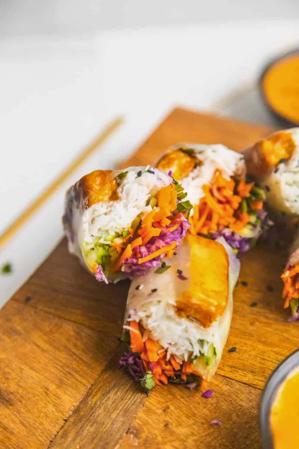 Fresh vegan spring rolls filled with rice noodles, shredded carrots, purple cabbage, cucumber, and fresh herbs, wrapped in translucent rice paper and cut in half to reveal the colorful filling. The rolls are arranged on a wooden board with dipping sauce nearby, showcasing a light and refreshing plant based meal.
